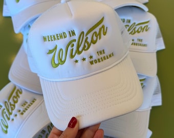 May include: A white trucker hat featuring a foam front panel and mesh back. The front is embroidered with the green text "WEEKEND IN Wilson" followed by three stars and the text "THE WORSHAMS" in a smaller green font, with a white cord trim.