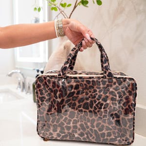 May include: A rectangular cosmetic bag with a leopard print pattern. The bag has a clear, glossy exterior and a zipper closure. The handles and trim are a matching leopard print. The bag is sitting on a white surface.