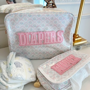 May include: A set of baby care accessories, including a diaper bag and a wipes case, both with a floral pattern in pink and blue. The diaper bag has the word "DIAPERS" in pink embroidery. A stack of diapers is also visible.