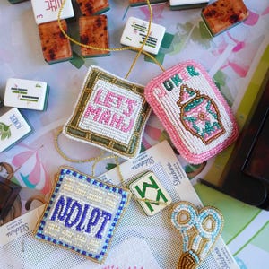 May include: Four beaded ornaments featuring Mahjong-inspired designs, including two square pieces with text reading LETS MAHJ and NOIPT, a pink rectangular piece with a vase graphic, and a pair of scissors, all with gold hanging cords.