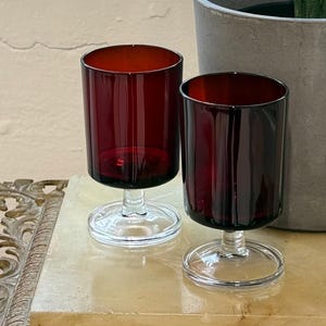 May include: Two burgundy-colored drinking glasses with clear stems. The glasses are cylindrical and sit on a decorative table with an ornate gold-colored frame. A potted plant with green leaves is in the background.