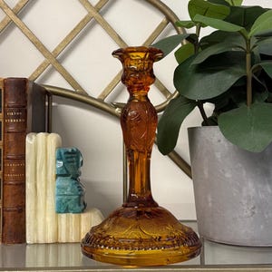 May include: Amber-colored glass candlestick holder with intricate floral and leaf patterns. The base and stem are detailed with embossed designs. A decorative piece for home decor.