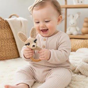 May include: A beige crochet baby rattle featuring a stuffed animal head with long ears, black embroidered eyes, and eyelashes attached to a smooth, round wooden ring. The toy is crafted with a textured yarn finish and a simple, neutral design.