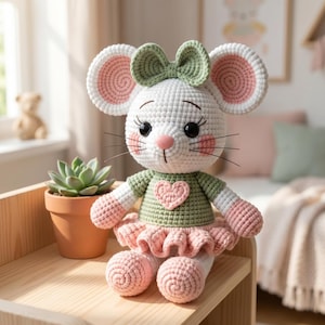 May include: A crocheted mouse doll featuring a white body, large ears with pink centers, and a sage green bow. The mouse wears a sage green shirt with a pink heart appliqué and a ruffled pink skirt, with pink accents on the cheeks and paws.