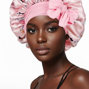 May include: A pink satin bonnet with a floral pattern and a large bow. The bonnet is designed to protect hair and features a soft, smooth texture. The bow is a solid pink color, adding a decorative element to the hair accessory.