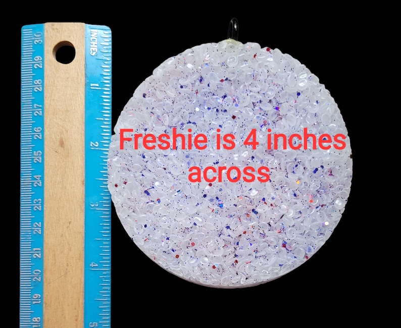 May include: A round, white air freshener with red and blue glitter, measuring 4 inches across. The text "Freshie is 4 inches across" is visible on the surface. A blue and wooden ruler is on the left side.