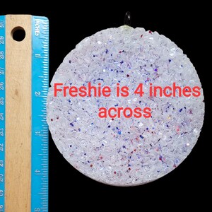 K&ouml;nnte beinhalten: Ein runder, wei&szlig;er Lufterfrischer mit roten und blauen Glitzer, etwa 10 cm im Durchmesser. Der Text "Freshie is 4 inches across" ist sichtbar. Ein blaues und h&ouml;lzernes Lineal befindet sich links.