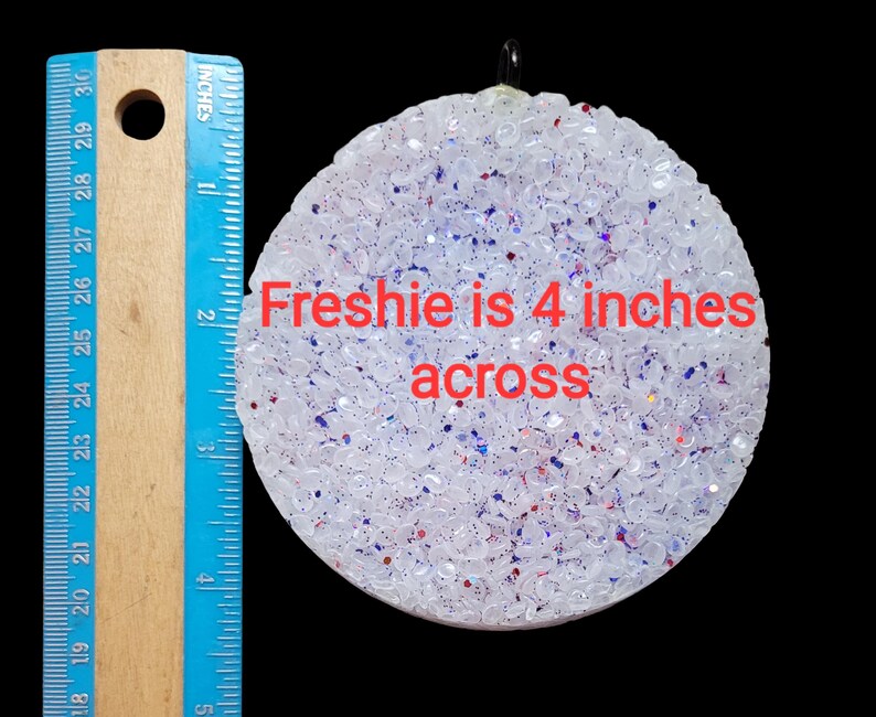 K&ouml;nnte beinhalten: Ein runder, wei&szlig;er Lufterfrischer mit roten und blauen Glitzer, etwa 10 cm im Durchmesser. Der Text "Freshie is 4 inches across" ist sichtbar. Ein blaues und h&ouml;lzernes Lineal befindet sich links.