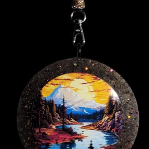 May include: A round pendant featuring a scenic landscape painting of a river, mountains, and a sunset. The pendant has a glittery, dark border and is attached to a black cord with decorative beads and a clasp.