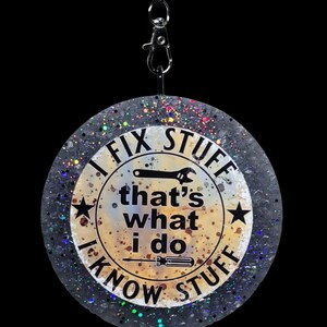 K&ouml;nnte beinhalten: Ein runder, glitzernder Anh&auml;nger mit dem Text "I FIX STUFF, that's what I do, I KNOW STUFF." Das Design beinhaltet einen Schraubenschl&uuml;ssel, einen Schraubendreher und Sterne. Er h&auml;ngt an einer schwarzen Kordel mit einem silbernen Verschluss.