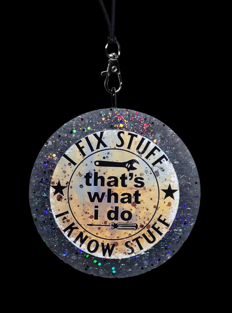K&ouml;nnte beinhalten: Ein runder, glitzernder Anh&auml;nger mit dem Text "I FIX STUFF, that's what I do, I KNOW STUFF." Das Design beinhaltet einen Schraubenschl&uuml;ssel, einen Schraubendreher und Sterne. Er h&auml;ngt an einer schwarzen Kordel mit einem silbernen Verschluss.