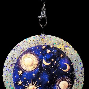 May include: A celestial-themed pendant with a starry night design. The pendant features a blue background with gold suns, moons, and stars. It is surrounded by a glittery, white border and attached to a beaded lanyard with a silver clasp.