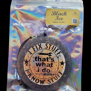 K&ouml;nnte beinhalten: Ein runder, schwarzer Lufterfrischer mit Glitzer und dem Text "I FIX STUFF, that's what I do, I KNOW STUFF." Der Lufterfrischer befindet sich in einer transparenten, irisierenden Verpackung mit einem "Black Ice"-Etikett.