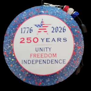 May include: A round, patriotic ornament with a blue glitter background and red and white accents. The center features the text "1776 2026 250 YEARS UNITY FREEDOM INDEPENDENCE" and a star with red and blue stripes.