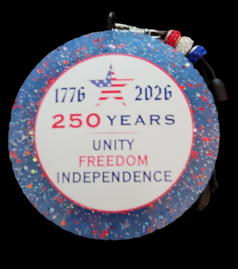 May include: A round, patriotic ornament with a blue glitter background and red and white accents. The center features the text "1776 2026 250 YEARS UNITY FREEDOM INDEPENDENCE" and a star with red and blue stripes.