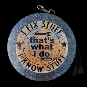 K&ouml;nnte beinhalten: Ein runder Schl&uuml;sselanh&auml;nger vor schwarzem Hintergrund. Er hat einen silbernen Verschluss und eine schwarze Kordel mit zwei schwarzen Quasten. In der Mitte steht der Text "I FIX STUFF that's what I do I KNOW STUFF" mit einer Schraubenschl&uuml;ssel- und Schraubendreher-Grafik.