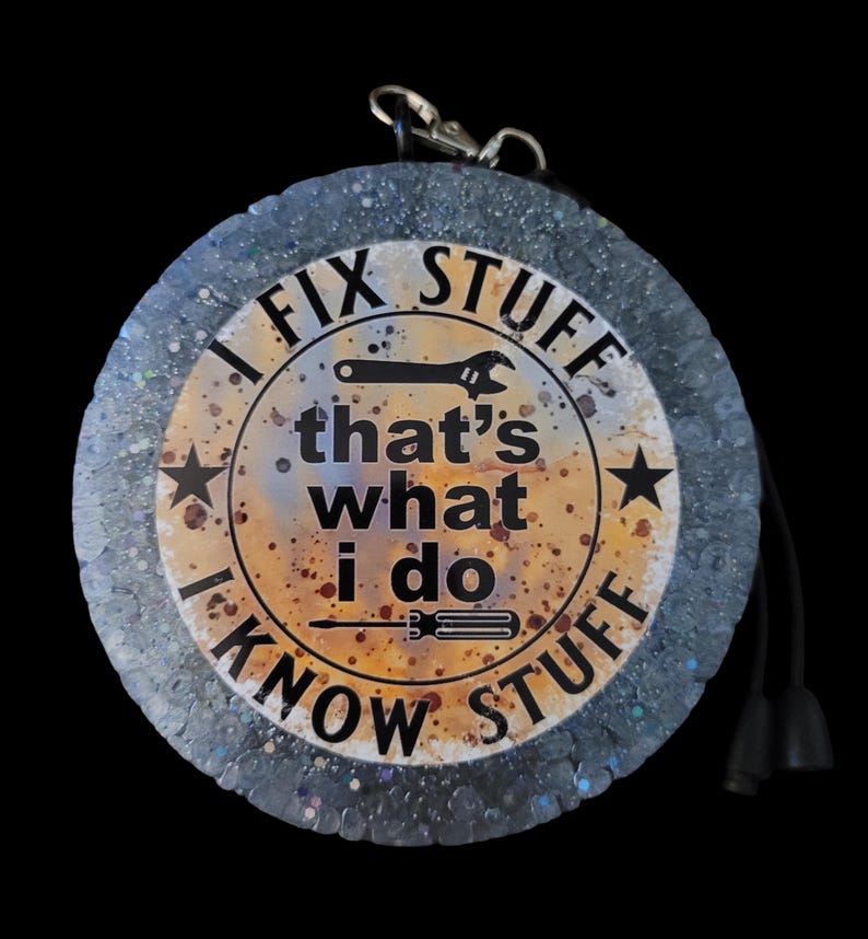 K&ouml;nnte beinhalten: Ein runder Schl&uuml;sselanh&auml;nger vor schwarzem Hintergrund. Er hat einen silbernen Verschluss und eine schwarze Kordel mit zwei schwarzen Quasten. In der Mitte steht der Text "I FIX STUFF that's what I do I KNOW STUFF" mit einer Schraubenschl&uuml;ssel- und Schraubendreher-Grafik.