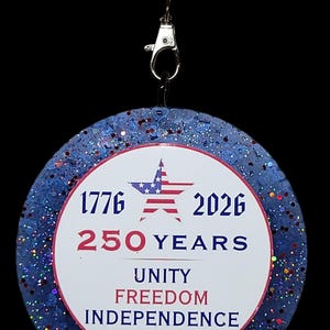 May include: A round, patriotic-themed pendant with a blue glitter border and red and white accents. The center features the text "1776 2026 250 YEARS UNITY FREEDOM INDEPENDENCE" and an American flag star. It hangs from a black cord with red, white, and blue beaded accents.
