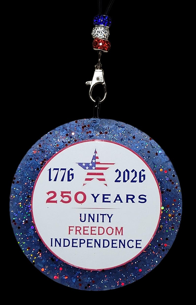May include: A round, patriotic-themed pendant with a blue glitter border and red and white accents. The center features the text "1776 2026 250 YEARS UNITY FREEDOM INDEPENDENCE" and an American flag star. It hangs from a black cord with red, white, and blue beaded accents.
