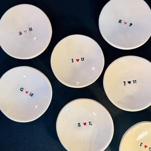 Peut inclure: Plusieurs petits plats en céramique blanc cassé. Chaque plat présente une initiale ou des initiales noires, avec un symbole de cœur rouge. Les plats sont disposés sur une surface sombre. Le texte comprend les initiales suivantes : B♥H, C♥M, E♥P, I♥U, J♥H et E♥K.