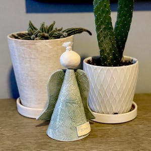 Puede incluir: Una figurita de ángel de cerámica con un diseño floral verde y blanco. El ángel tiene una cabeza blanca y un cuerpo cónico verde. El ángel sostiene una pequeña etiqueta blanca que dice "Mamá".