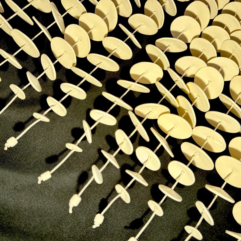 May include: A collection of multiple vertical strands featuring small, pale cream-colored circular discs strung along thin cords. Each strand is finished with a small knot at the bottom, creating a repetitive, linear pattern of light-toned shapes.