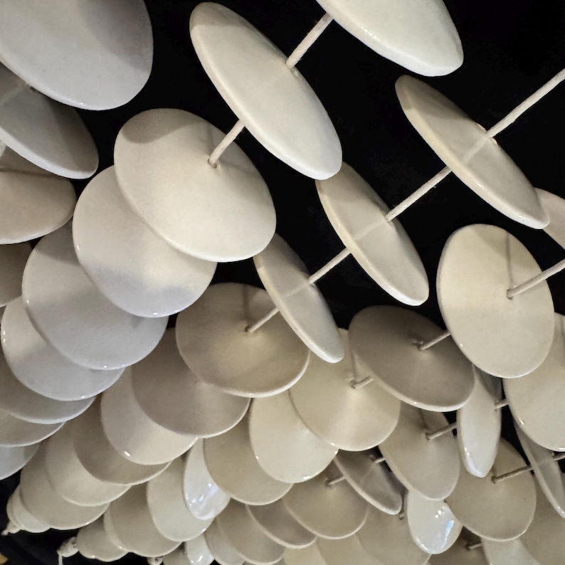 May include: A collection of white circular ceramic discs suspended on thin vertical rods. The discs are arranged in a repeating grid pattern against a dark background, creating a structured display of multiple smooth, round, off-white elements.