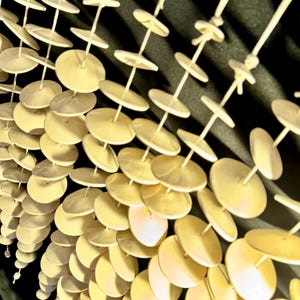 May include: A decorative hanging mobile composed of multiple vertical strands. Each strand features numerous small, pale yellow, circular discs spaced evenly along a thin, rigid rod, creating a repetitive, layered, and geometric visual pattern.