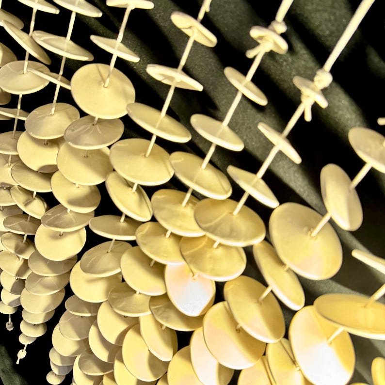 May include: A decorative hanging mobile composed of multiple vertical strands. Each strand features numerous small, pale yellow, circular discs spaced evenly along a thin, rigid rod, creating a repetitive, layered, and geometric visual pattern.