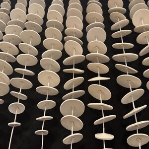May include: Multiple vertical strands of white circular ceramic discs strung together on thin white cord. Each strand features numerous flat, round, matte-finished clay pieces spaced evenly along the length of the string, creating a repetitive pattern.