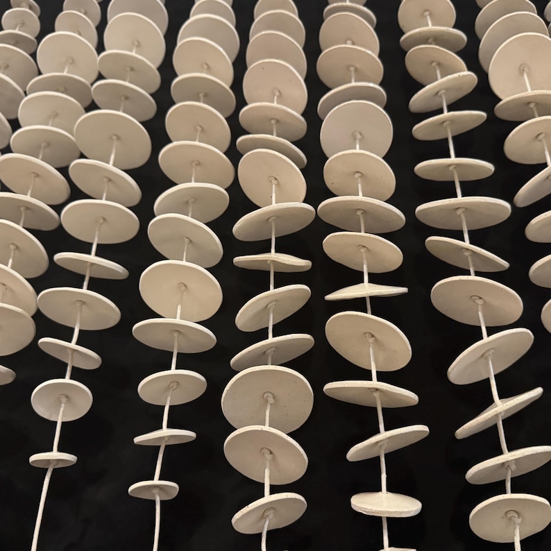 May include: Multiple vertical strands of white circular ceramic discs strung together on thin white cord. Each strand features numerous flat, round, matte-finished clay pieces spaced evenly along the length of the string, creating a repetitive pattern.