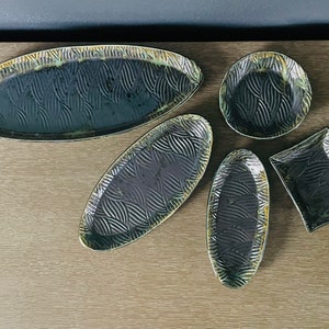 May include: A collection of five ceramic serving dishes in various shapes: an oval platter, two oval trays, a round plate, and a square dish. Each piece features a dark teal glaze with a textured leaf pattern and a gold-toned rim.