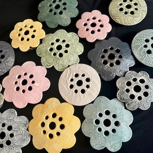 May include: A collection of ceramic flower frogs in various colors including pink, yellow, green, grey, and cream. Each piece features a scalloped edge, a circular shape, and multiple holes for arranging stems, with textured leaf patterns on the surface.
