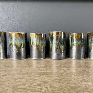 May include: A set of six cylindrical ceramic tumblers featuring a two-tone glaze. The upper portion displays a dripped, variegated pattern of ochre, teal, and dark brown, while the lower portion is finished in a smooth, matte charcoal gray.