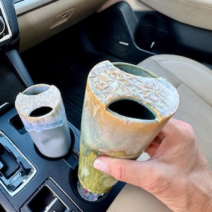 May include: Two ceramic vases with a unique design. One is held in a hand, the other sits in a car cup holder. The vases have a textured floral pattern around the top and a color gradient of blue, green, and gold.