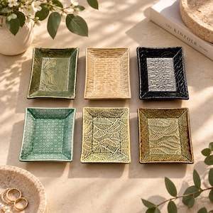 May include: Six rectangular ceramic trinket trays arranged in two rows. Each tray features a unique embossed pattern, including floral, leaf, and geometric designs in various colors such as green, beige, black, and gold, with raised edges.