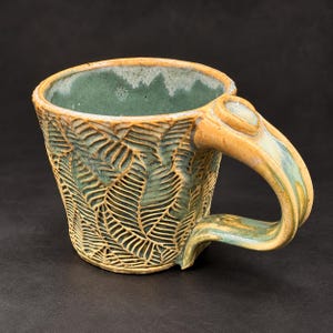 Puede incluir: Una taza de cerámica con un exterior texturizado que presenta un patrón de hojas de helecho en relieve en tonos verde salvia y amarillo dorado. El interior está esmaltado en verde salvia liso y el asa tiene un acabado de esmalte amarillo brillante.