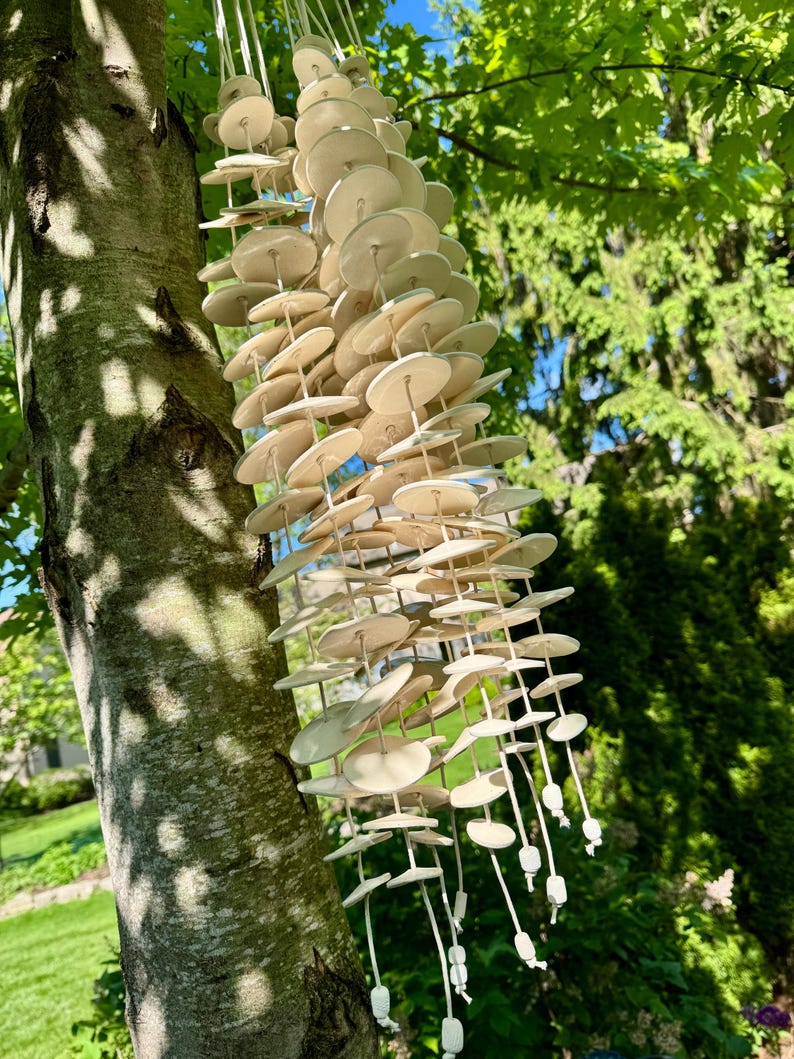 May include: A hanging wind chime composed of numerous off-white, circular ceramic discs strung together on multiple vertical cords. The discs are arranged in several cascading strands, each finished with a small, square ceramic bead at the bottom.