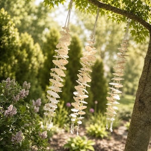 May include: Three hanging wind chimes made of numerous small, circular, off-white ceramic discs strung together on thin white cords. The chimes are suspended from a tree branch, featuring a vertical arrangement of flat, round pieces.