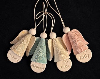 May include: Four ceramic angel ornaments with textured wings and cone-shaped bodies in beige, blue-green, and pink. Each angel features a round base stamped with the names Robert, Michael, I.D., or Lilly, and includes a hanging cord.