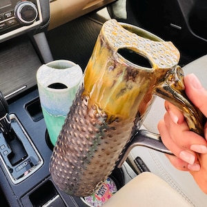 May include: Two ceramic tumblers with lids. One is a tall, textured brown and amber glazed mug with a handle. The other is a smooth, light green and white ombre tumbler. Both feature a distinct top opening design for drinking, held in a car interior.