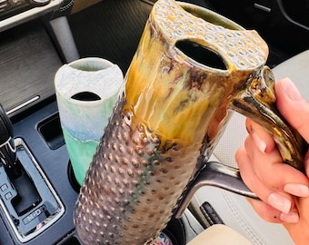 Può includere: Un'alta tazza cilindrica in ceramica con una superficie ruvida e punteggiata e una smaltatura lucida marrone e ambra, dotata di manico e coperchio specializzato. Accanto si trova un bicchiere in ceramica più piccolo, verde chiaro e bianco.
