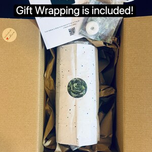 May include: A cardboard shipping box containing a cylindrical item wrapped in white paper with colorful confetti dots and a circular succulent sticker. The box includes brown packing paper, a white card, and a small white organza gift bag.