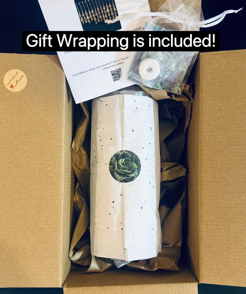 May include: A cardboard shipping box containing a cylindrical item wrapped in white paper with colorful confetti dots and a circular succulent sticker. The box includes brown packing paper, a white card, and a small white organza gift bag.