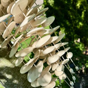 May include: A wind chime featuring numerous smooth, off-white circular ceramic discs suspended on thin, light-colored strings. The discs are arranged in multiple vertical strands, creating a layered, cascading structure that hangs from a central point.