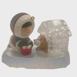 Op de afbeelding: Een beeldje van een kind in een parka, een hond en een iglo. Het kind houdt een rode emmer vast met het nummer "1999". De hond is bruin en wit, en de iglo is doorschijnend.