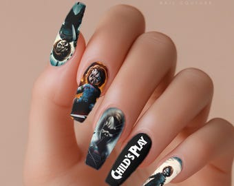 May include: A hand displaying long, coffin-shaped nail art. The nails feature images of the Chucky doll from the Child's Play horror film series. The nail art includes images of Chucky, the film's title, and other related graphics.