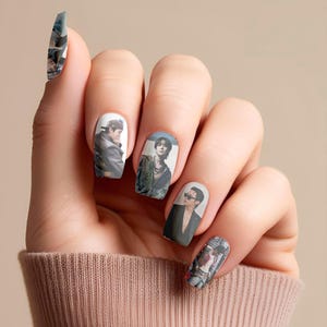 May include: A hand with painted fingernails. Each nail features a different image of a person. The nail art includes images of people in various outfits, including a leather jacket, sunglasses, and a suit. The nail polish is white.
