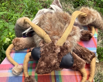 May include: A plush insect toy with brown fur, black eyes, and tan legs and antennae. The toy is lying on a colorful plaid fabric, with green foliage in the background.