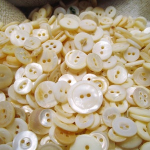 May include: A collection of off-white buttons with various shapes and sizes. The buttons have two or four holes and a slightly iridescent sheen. The buttons are scattered, creating a textured surface.